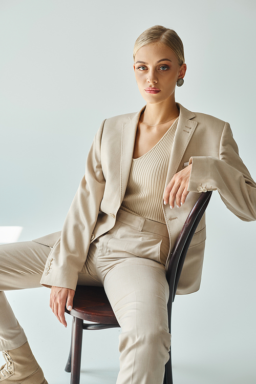 blonde fashion model in pastel beige suit sitting on chair and looking at camera on grey backdrop