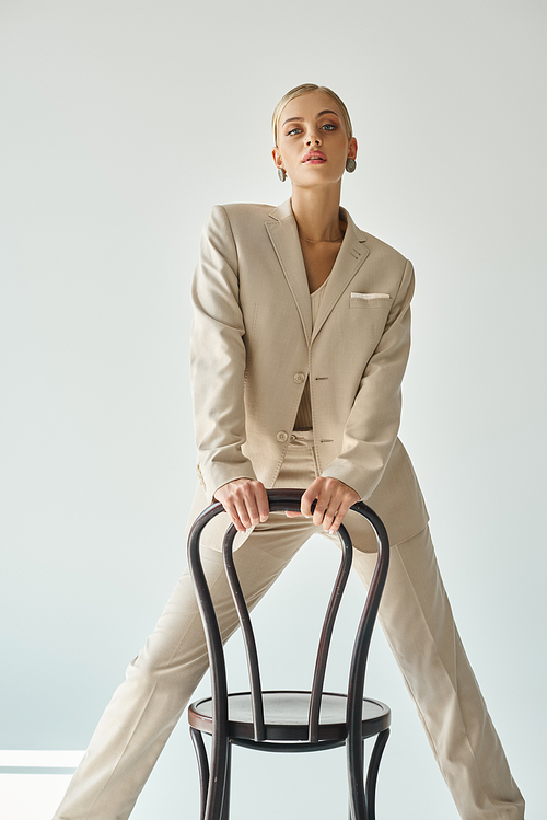 alluring fashion model in pastel beige suit posing with chair and looking at camera on grey backdrop