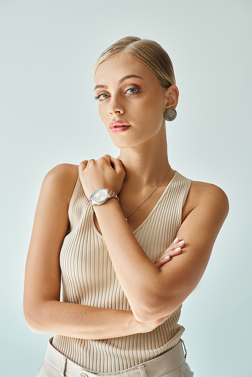 portrait of pretty blonde woman in beige tank top and wristwatch looking at camera on grey