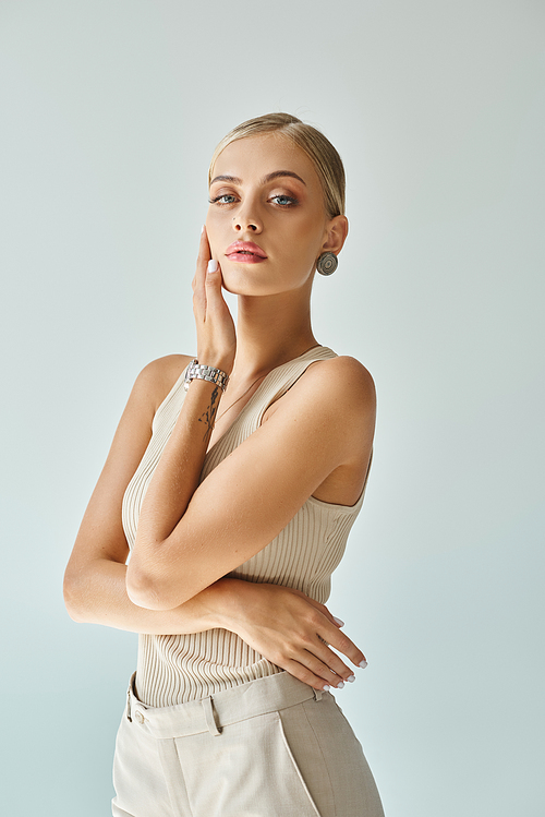 tattooed woman in beige tank top and wristwatch touching face with natural makeup on grey backdrop