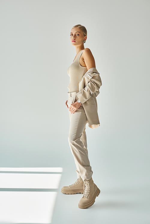full length of fashionable woman in total beige casual attire standing on grey backdrop in sunlight