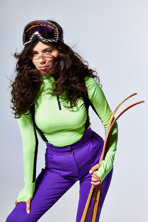 brunette pretty woman with curly hair posing in trendy sporty clothes and holding skis on grey