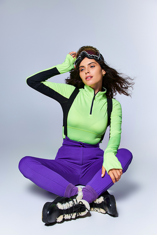 sporty young woman with curly hair posing in trendy active wear while sitting on grey backdrop