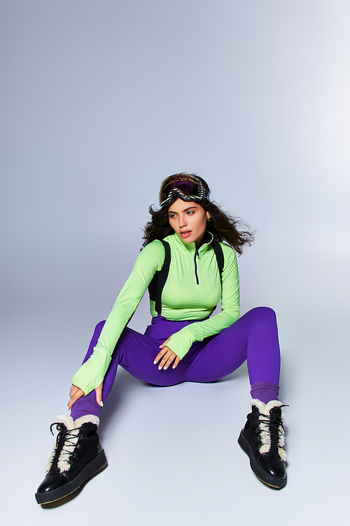 brunette sporty woman with curly hair posing in trendy active wear while sitting on grey backdrop