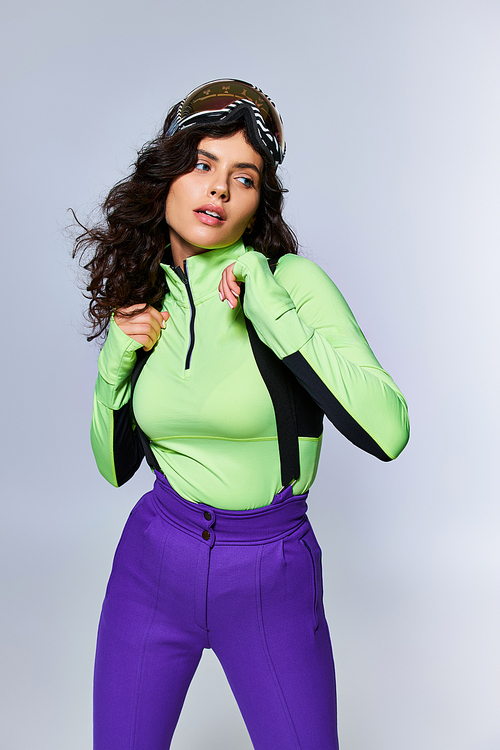 winter sport, sporty brunette woman with curly hair posing in ski clothes on grey backdrop