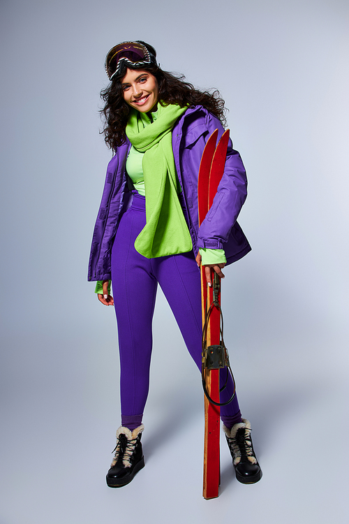 winter sport, cheerful woman with curly hair posing in active wear with puffer jacket and skis