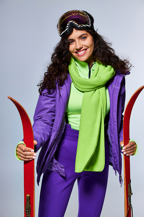 winter leisure, positive woman with curly hair posing in active wear with puffer jacket and skis