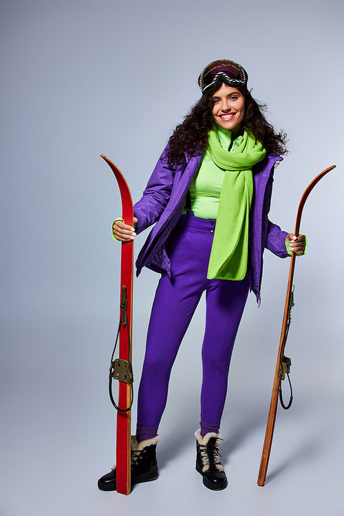 winter sport, smiling woman with curly hair posing in active wear with puffer jacket and skis