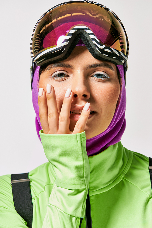 young woman in winter ski clothes with balaclava on head posing with hands near face on grey