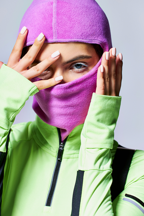 portrait of woman with blue eyes wearing purple ski mask and looking at camera on grey backdrop