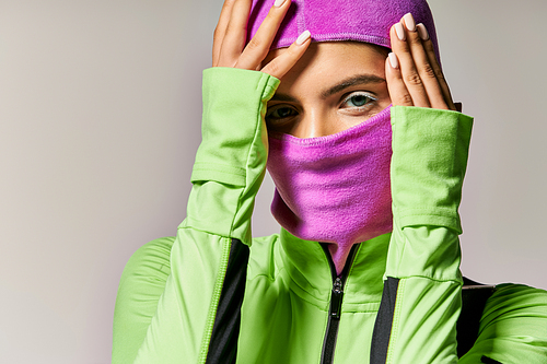 portrait of sportive woman with blue eyes wearing purple ski mask and looking at camera on grey