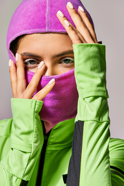 portrait of sportive woman with blue eyes wearing ski mask and looking at camera on grey backdrop