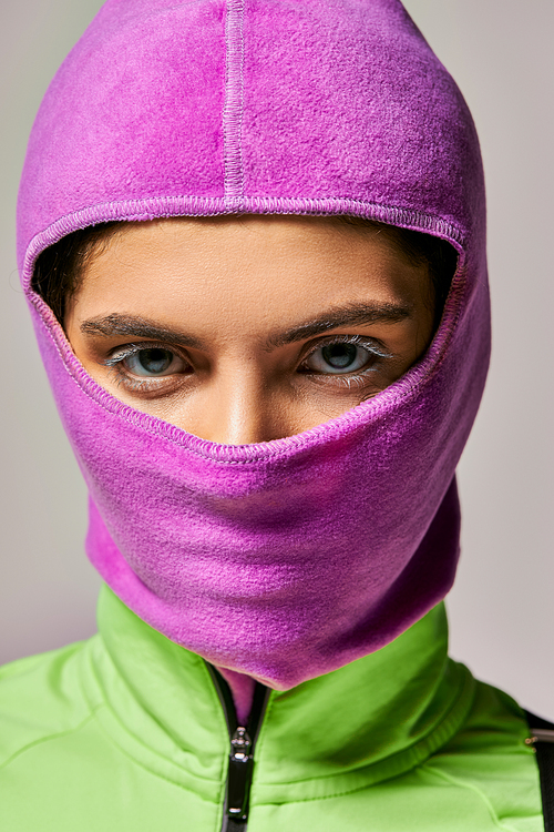 portrait of young woman with blue eyes in purple ski mask looking at camera on grey backdrop