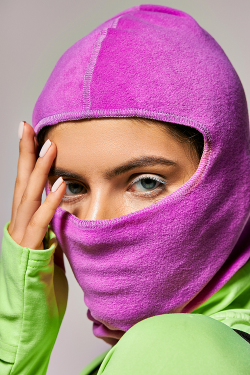 portrait of woman with blue eyes in purple ski mask posing with hand near face on grey backdrop