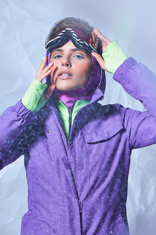 beautiful woman in balaclava and winter jacket wearing ski googles on grey backdrop, snowy day