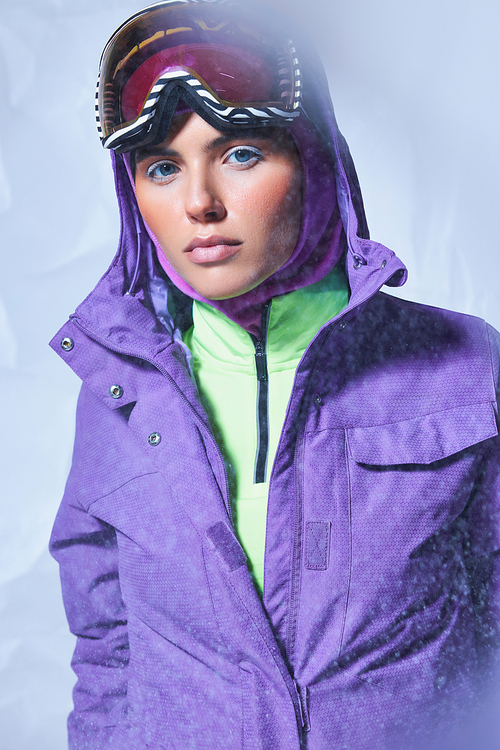 attractive woman in balaclava, purple winter jacket and ski googles posing on grey, snowing day