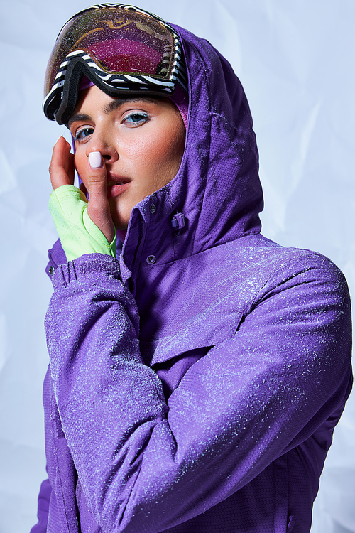 attractive woman in balaclava and ski googles on grey background, snow on purple winter jacket