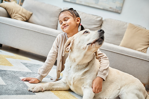 pleased girl sitting on carpet and stroking labrador in modern living room, kid and furry friend