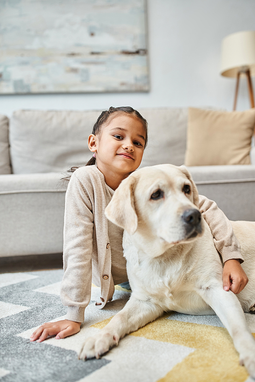 cute girl sitting on carpet and stroking labrador in modern living room, kid and furry friend