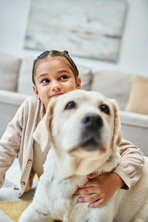 happy cute girl sitting on carpet and stroking dog in modern living room, kid and labrador