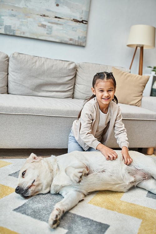 cheerful girl in casual attire smiling and stroking dog in modern living room, kid and labrador