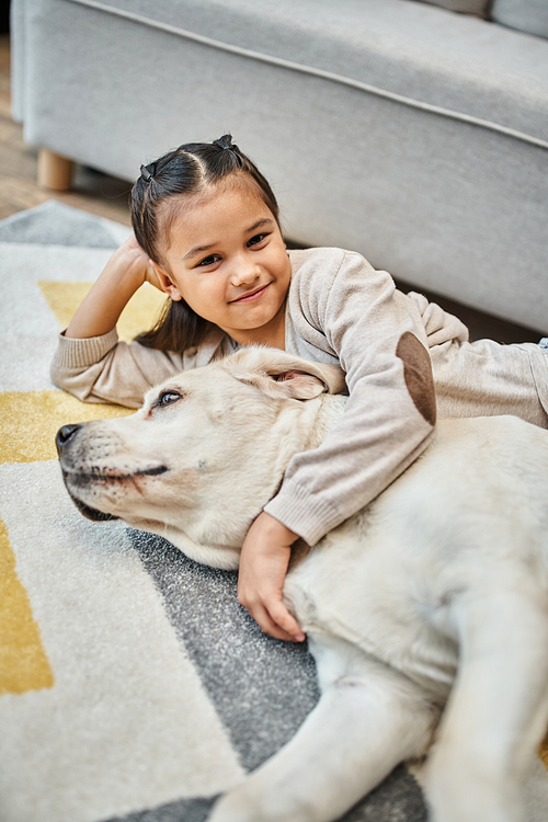 positive girl in casual attire smiling and stroking dog in modern living room, child and labrador