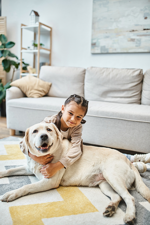 cheerful girl in casual attire smiling and stroking dog in modern living room, child and labrador