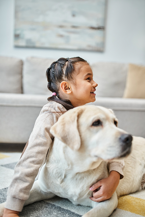 joyful kid in casual attire smiling and stroking dog in modern living room, child and labrador