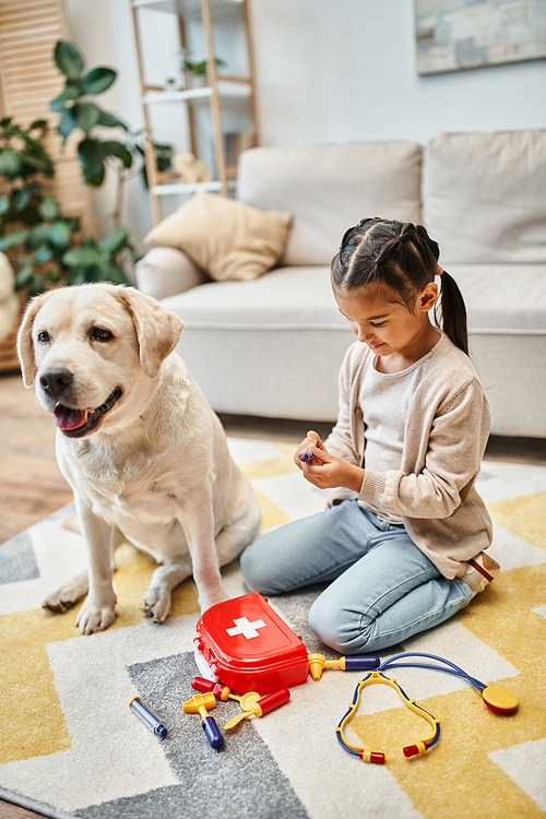 cheerful kid in casual attire playing doctor with labrador in modern living room, toy first aid kit