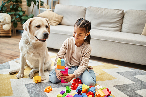 smiling girl playing with colorful toy blocks near labrador in living room, building tower game