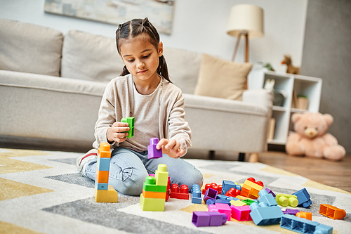 cute little girl playing with colorful toy blocks on carpet in living room, building tower game