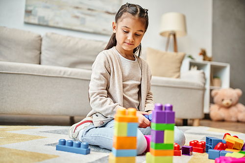 cute girl playing with colorful toy blocks on carpet in living room, building tower game