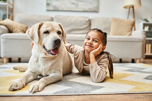 happy elementary age girl in casual attire lying on carpet with labrador in modern living room