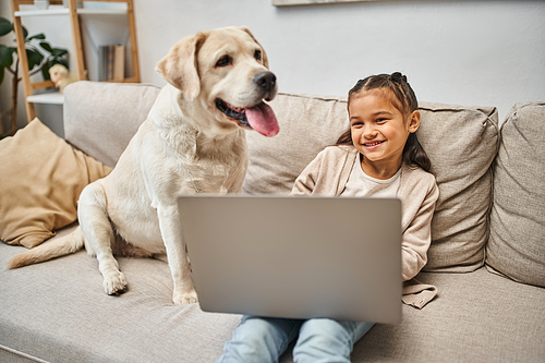 happy elementary age kid sitting on sofa and using laptop near labrador dog in modern living room