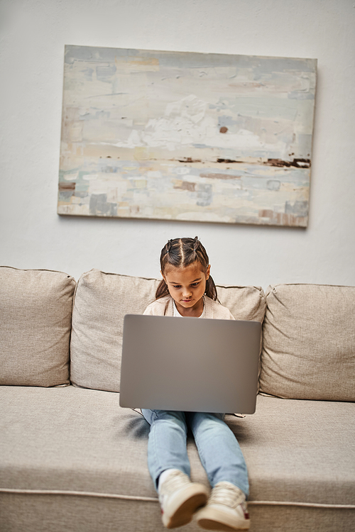 happy elementary age girl sitting on sofa and using laptop in modern living room, e-learning