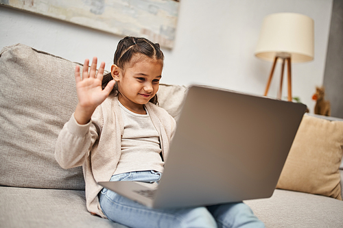 happy elementary age girl sitting on sofa and waving hand at laptop in living room, online lesson