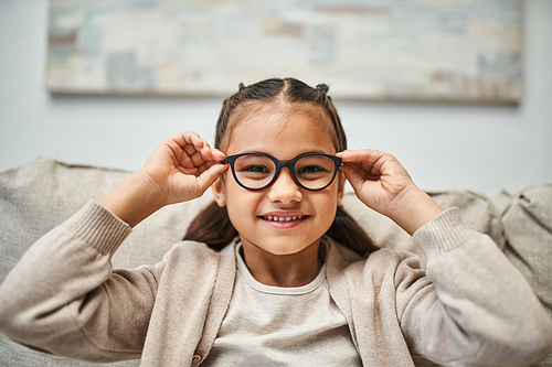 happy elementary age girl in casual attire wearing eyeglasses and looking at camera in living room