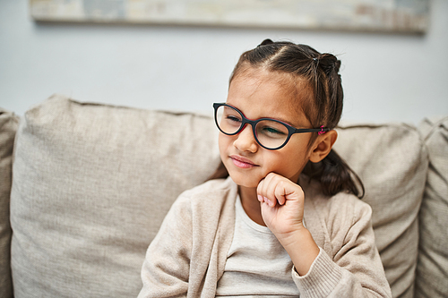 pensive elementary age girl in casual attire wearing eyeglasses and looking away in living room