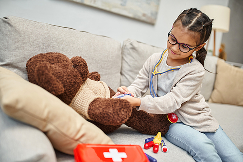 happy kid in casual wear and eyeglasses playing doctor with teddy bear on sofa in living room