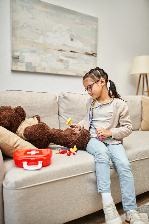 happy kid in casual wear and eyeglasses playing doctor with soft teddy bear on sofa in living room