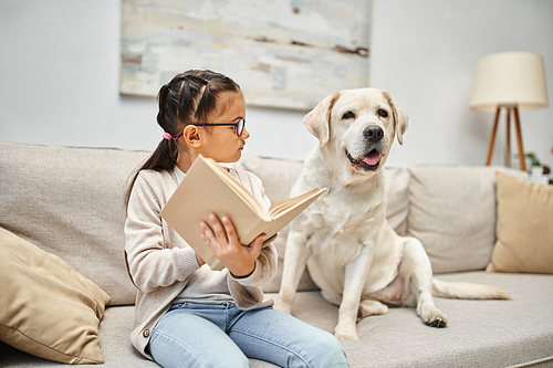 smart girl in casual wear and eyeglasses reading book to labrador dog on sofa in living room