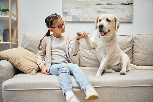 happy elementary age girl in eyeglasses holding paw of labrador and sitting on sofa in living room