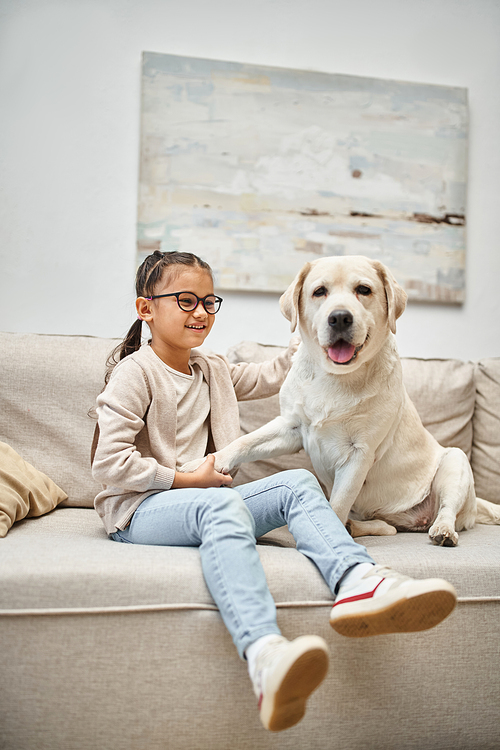 happy elementary age girl in eyeglasses holding paw of labrador and sitting on sofa in living room