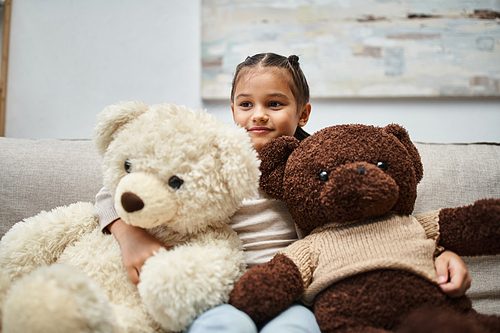 cute elementary age child in casual wear sitting on sofa with soft teddy bears in living room