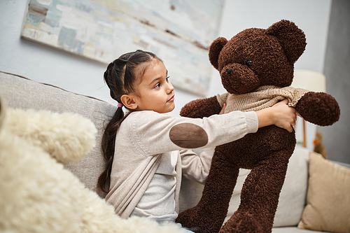 cute elementary age kid in casual wear sitting on sofa with soft teddy bears in living room