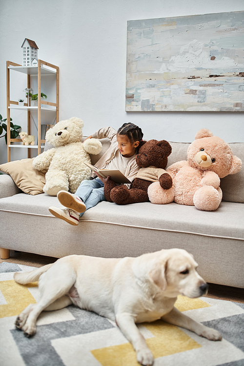 adorable girl sitting on sofa with soft teddy bears and reading book near labrador in living room