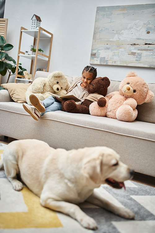 adorable girl sitting on sofa with soft teddy bears and reading book near labrador on carpet