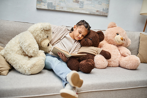 adorable girl sitting on sofa with soft teddy bears and reading book in modern living room