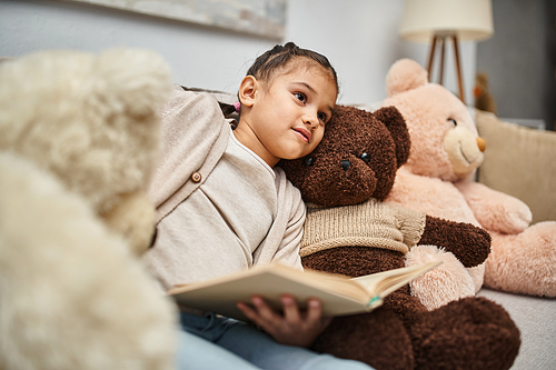 cute girl sitting on comfortable sofa with soft teddy bears and reading book in modern living room