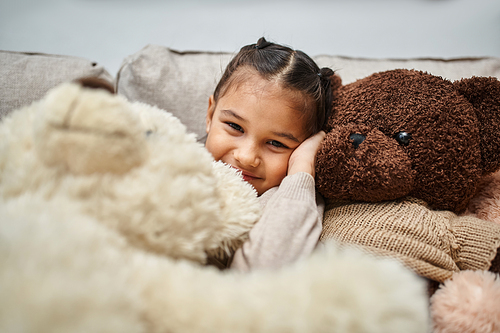 cheerful elementary age girl sitting among soft teddy bears on couch in modern living room